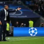 A focused Vincent Kompany gesturing on the touchline during a previous Champions League match, with the iconic UCL logo in the background.