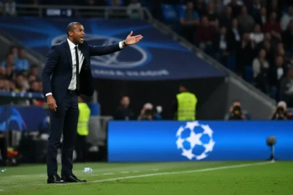 A focused Vincent Kompany gesturing on the touchline during a previous Champions League match, with the iconic UCL logo in the background.